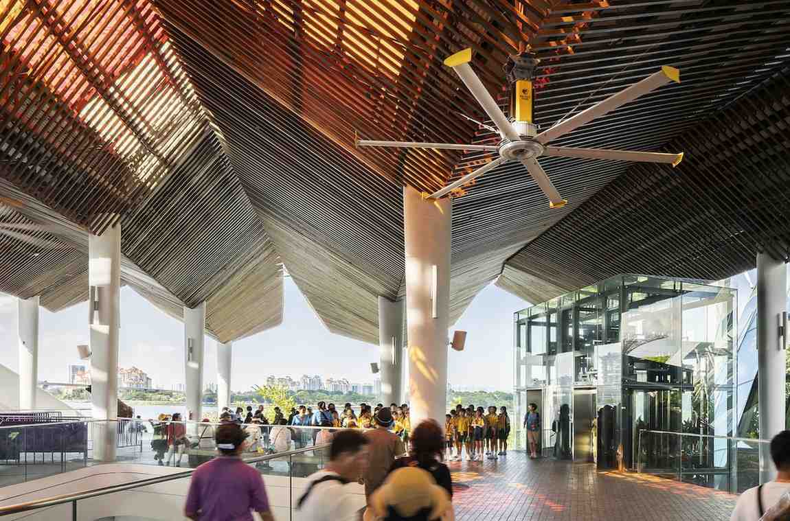 A group of individuals strolls inside a vast building with a prominent ceiling fan.